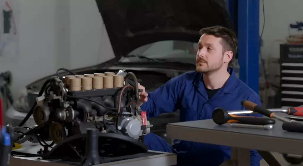A realistic horizontal photo of a skilled mechanic in a well-lit automotive workshop, focused on repairing a Geo engine. The mechanic, wearing a blue coveralls and a white safety helmet, is kneeling beside an open engine bay of a Geo vehicle, with a variety of automotive tools (wrenches, socket set, torque wrench) neatly arranged on a metal workbench. The engine is partially disassembled, showing visible pistons, crankshaft, and timing belt. The background includes a clean, organized workshop setting with a subtle hint of a Geo car chassis and a toolbox. The lighting highlights the metallic surfaces and the mechanic’s concentration, creating a professional and inviting atmosphere.
