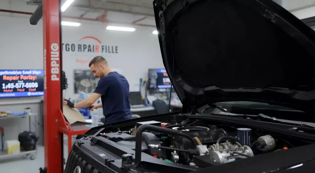 A high-resolution horizontal photo of a professional automotive repair shop interior, featuring a mechanic in a clean work environment. The scene shows a large, open engine bay of a car on a hydraulic lift, with a variety of tools and parts neatly arranged on a workbench. The focus is on the engine, with visible pistons, crankshaft, and oil filter, illuminated by bright overhead lights. The background includes a branded repair shop sign and a phone number displayed on a digital screen, but no visible text or numbers in the image itself. The overall composition conveys expertise and precision in engine overhaul.