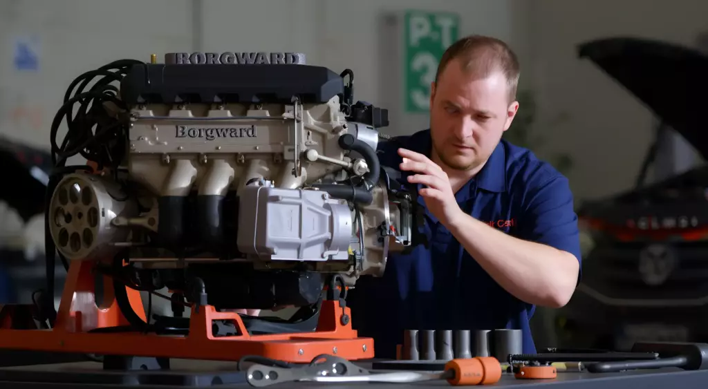 A realistic, high-resolution horizontal photo of a professional automotive repair workshop. In the foreground, a mechanic in a clean uniform is carefully inspecting a Borgward engine block placed on a hydraulic lift. The workbench is organized with various tools (wrenches, sockets, torque wrench) neatly arranged. The background shows a garage setting with subtle safety signage and a clean, well-lit environment. The lighting is bright and evenly distributed, emphasizing the metallic surfaces and the mechanic’s focused expression.
