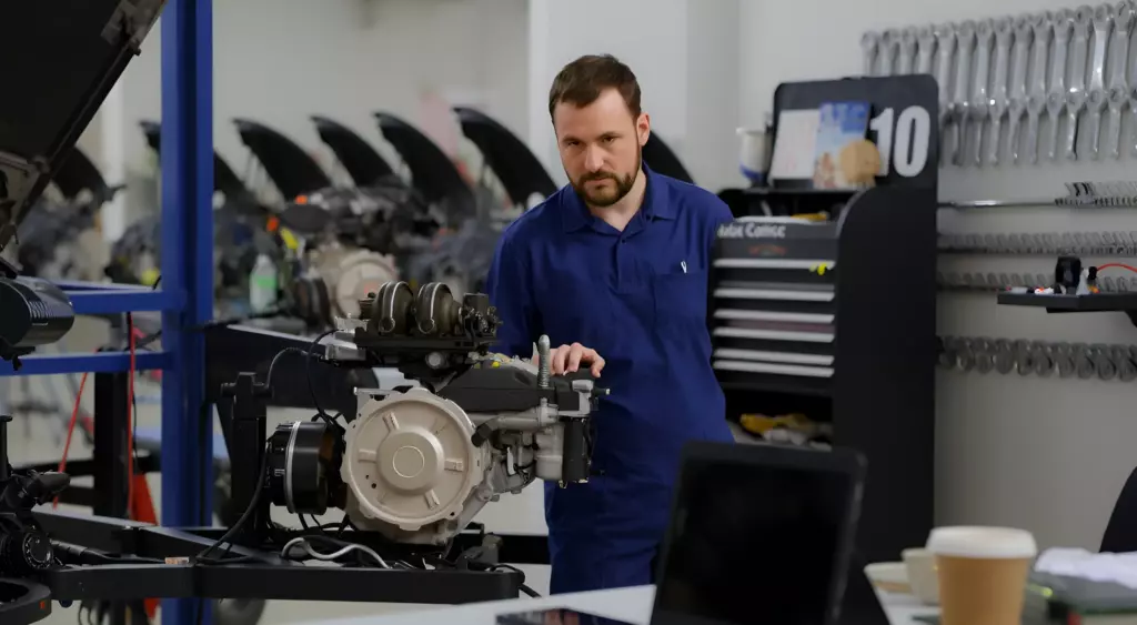 A wide, horizontal photo of a modern automotive repair workshop. In the foreground, a mechanic in a blue jumpsuit is inspecting a car engine on a hydraulic lift. The engine is partially disassembled, with visible pistons and crankshaft. In the background, other engines are on lifts and a wall-mounted toolbox holds various wrenches and sockets. A phone sits on a desk next to a cup of coffee, its screen off. The lighting is bright and even, highlighting the metallic surfaces and the clean, organized environment. No text, letters, or digits should appear in the image.