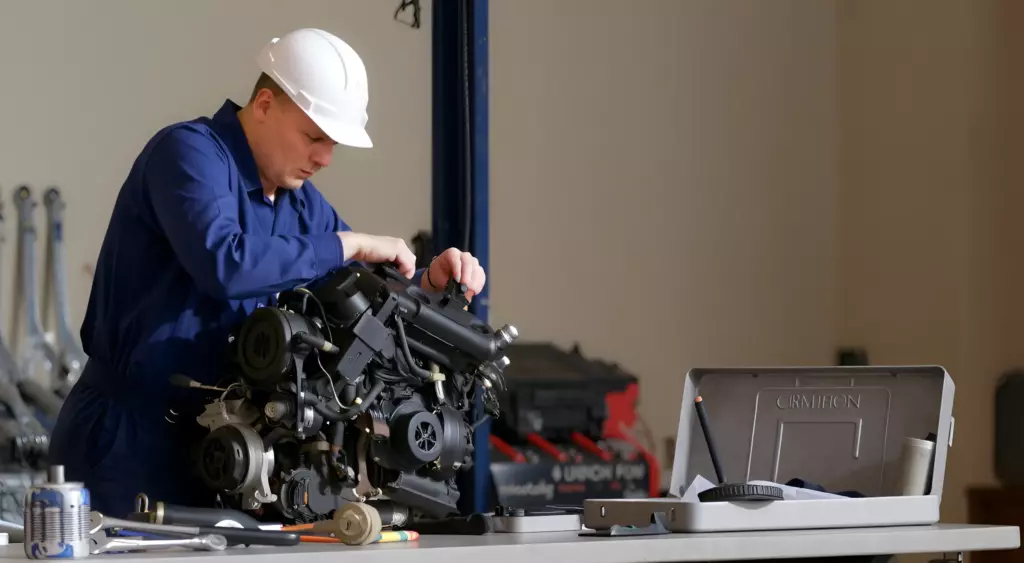 A realistic photo of a mechanic in a workshop, working on a car engine mounted on a lift, surrounded by tools and engine parts, with a phone on a nearby desk, all depicted in a horizontal format