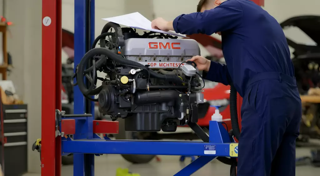A realistic horizontal photo of a professional automotive repair shop interior, featuring a mechanic in blue coveralls working on a GMC engine mounted on a hydraulic lift. The scene includes organized tools, engine components, and a visible service manual, all set in a clean, well-lit environment with a subtle background of other vehicle parts.