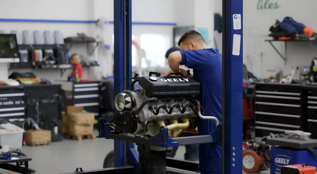 A professional automotive repair workshop scene featuring a mechanic in blue coveralls working on a Geely engine on a hydraulic lift, surrounded by organized tools and parts, with a clean, well-lit environment