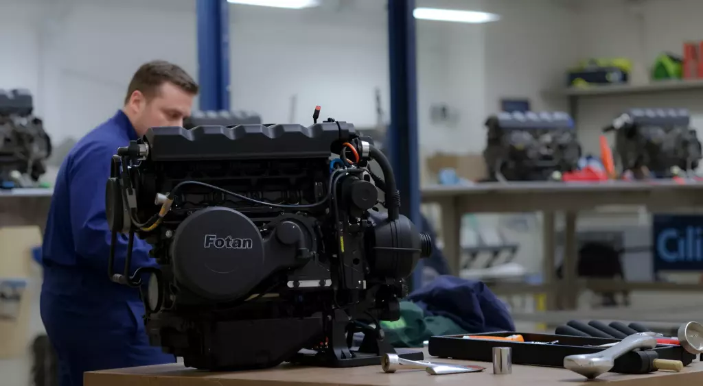 A realistic photo of a professional automotive repair workshop. In the foreground, a mechanic in a blue coverall is kneeling beside a large, partially disassembled internal combustion engine. The engine has a subtle, visible Foton logo on its side. The workshop is well-lit with overhead lights, and there are various automotive tools (wrenches, torque wrench, socket set) neatly arranged on a metal workbench. The background shows a clean, organized garage environment with a few other engine parts and a workbench table. The scene conveys expertise, precision, and a focus on engine repair.