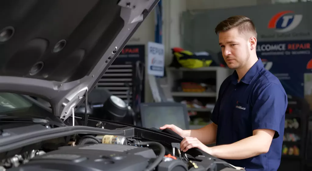 A high-resolution, horizontally oriented photo of a professional mechanic in a clean, well-lit automotive repair shop, focused on a car engine disassembly. The scene includes a detailed Brilliance engine block, various engine components (pistons, camshaft, timing belt), a set of precision tools (torque wrench, socket set, screwdriver) neatly arranged on a workbench, and a safety mask and gloves on the mechanic’s hands. The background shows a neatly organized workshop with a subtle industrial feel, emphasizing craftsmanship and technical expertise.